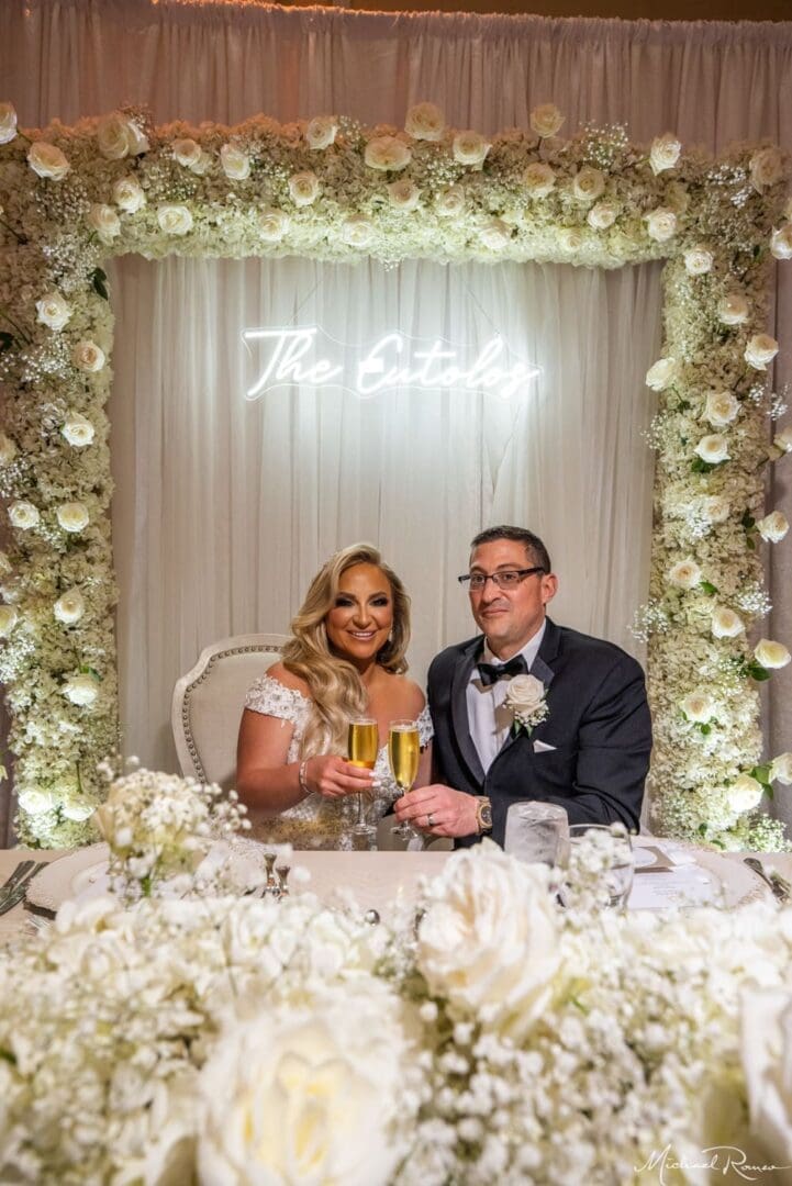 A bride and groom sitting at a table with flowers in the background.