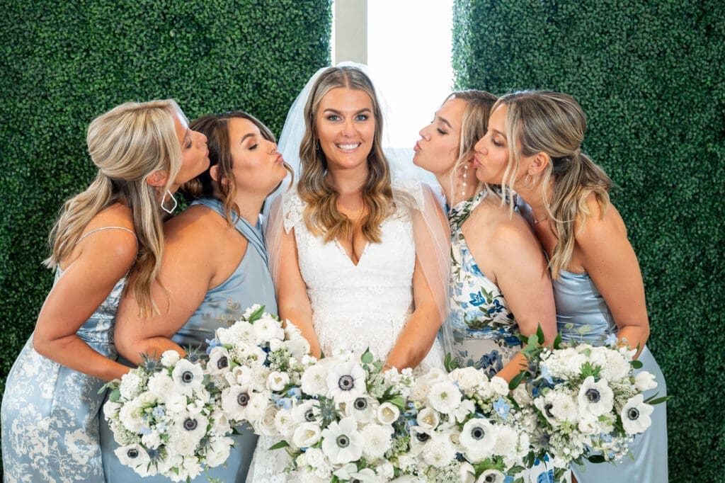 A bride and her bridesmaids kiss in front of a green wall.